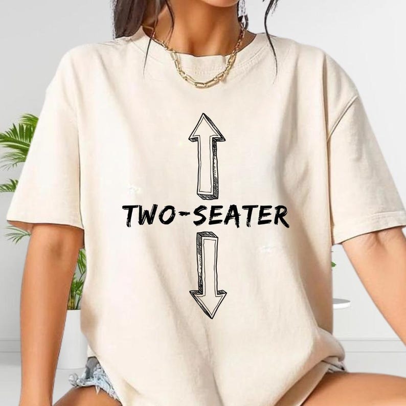 A person wearing a white t-shirt with a graphic design featuring two arrows pointing in opposite directions, with the text "TWO-SEATER" below the arrows.