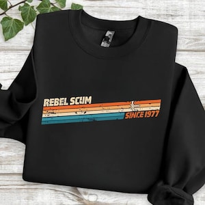 May include: Black sweatshirt with the text "REBEL SCUM" above a retro design of orange, tan, and blue stripes, with "SINCE 1977" in orange. The sweatshirt has a crew neck and long sleeves.