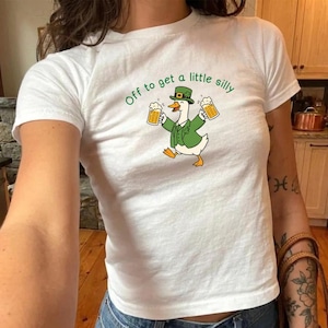 May include: White t-shirt with a cartoon goose wearing a green hat and jacket, holding two mugs of beer. The text "Off to get a little silly" is arched above the goose in green letters.