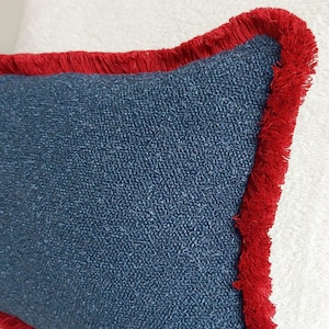 Könnte beinhalten: Nahaufnahme eines dekorativen Kissens mit einem strukturierten marineblauen Stoff und einem leuchtend roten Fransenrand. Die Form des Kissens ist leicht unregelmäßig, was zu seinem einzigartigen Design beiträgt. Die rote Franse bildet einen auffälligen Kontrast.