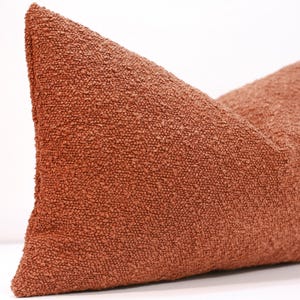 Può includere: Un cuscino rettangolare in tessuto bouclé testurizzato e granuloso di colore arancione terracotta uniforme. Il cuscino presenta un angolo pronunciato e una consistenza superficiale densa e irregolare creata dalla lavorazione a ricciolo del filato.