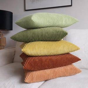 Op de afbeelding: Een stapel van vijf rechthoekige sierkussens met een textuur van corduroy stof. De kussens zijn in een verticale kolom gerangschikt en tonen een kleurverloop van boven naar beneden: lichtgroen, olijfgroen, mosterdgeel, roestoranje en perzik.