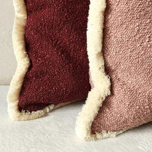 Könnte beinhalten: Nahaufnahme von zwei strukturierten Zierkissen. Eines ist tief burgunderfarben, das andere zartrosa. Beide Kissen haben einen cremefarbenen Fransenbesatz. Die Kissen sind wahrscheinlich für die Heimdekoration.