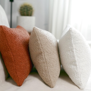Könnte beinhalten: Drei strukturierte Zierkissen in Orange-, Beige- und Weißtönen. Die Kissen haben eine weiche, noppenartige Textur und sind auf einer hellen Oberfläche angeordnet. Diese Kissen sind ein dekoratives Element für einen Wohnraum.