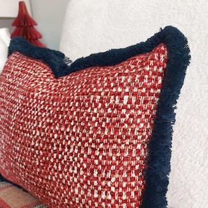 May include: Close-up of a decorative pillow with a red and white woven pattern. The pillow has a dark blue fringe trim around the edges. The pillow is on a white surface.