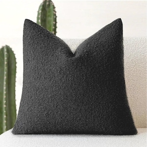 Könnte beinhalten: Ein quadratisches, strukturiertes schwarzes Kissen mit einer spitzen Oberseite. Das Kissen besteht aus einem gewebten Stoff und liegt auf einer weißen Oberfläche. Im Hintergrund sind zwei grüne Kakteen zu sehen.