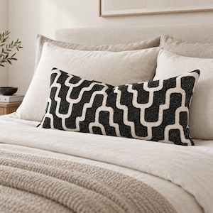 Könnte beinhalten: Ein rechteckiges Zierkissen mit einem schwarzen und cremefarbenen, strukturierten geometrischen Muster aus sich wiederholenden U-förmigen Linien. Das Kissen liegt horizontal auf einem Bett mit neutralfarbener Bettwäsche, beigen Kissen und einer Decke.