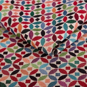 Puede incluir: Primer plano de una tela doblada con un patrón repetido de formas florales coloridas y estilizadas. El diseño presenta elementos rojos, naranjas, verdes, azules y morados sobre un fondo crema. La tela parece estar tejida.