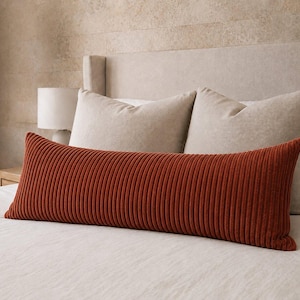 Peut inclure: Un long coussin lombaire rectangulaire présentant une couleur rouille profonde et une texture en velours côtelé vertical. Le coussin est placé horizontalement devant deux grands coussins carrés de couleur neutre sur un lit.