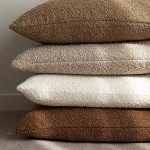 Peut inclure: Une pile de quatre coussins décoratifs texturés dans des tons neutres. Les coussins sont empilés les uns sur les autres, présentant des nuances de marron, beige et blanc. Le tissu a un aspect noueux et tissé.