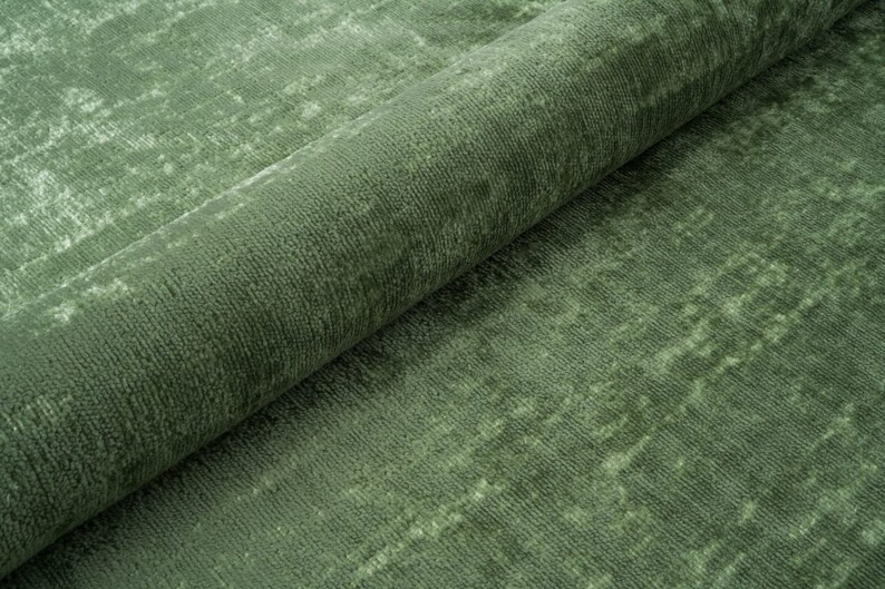 Puede incluir: Primer plano de una alfombra verde con un aspecto texturizado y desgastado. La alfombra est&aacute; doblada por la mitad, mostrando la textura y el color de la tela.