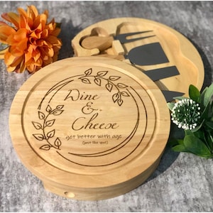 Puede incluir: Una tabla de queso redonda de madera con las palabras "Wine & Cheese" y la frase "get better with age (just like you!)" grabadas. La tabla tiene un diseño de hojas decorativas e incluye cuchillos para queso.