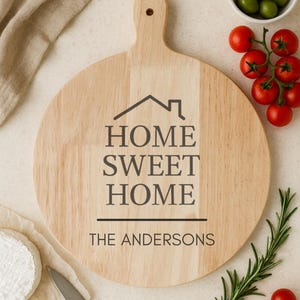 May include: A circular wooden chopping board with a handle, engraved with "HOME SWEET HOME" and "THE ANDERSONS". The board is surrounded by olives, tomatoes, rosemary, a knife, and cheese. Ideal for serving food in the kitchen.