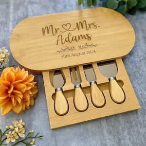 May include: An oval bamboo cheese board with a slide-out drawer containing four stainless steel cheese knives with matching bamboo handles. The board is engraved with the text Mr. & Mrs. Adams and the date 28th August 2024 with floral accents.