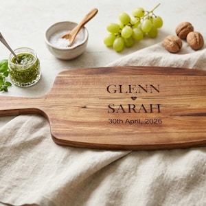 May include: A wooden chopping board with a handle, engraved with the names "GLENN" and "SARAH" and the date "30th April, 2026". The board is surrounded by grapes, walnuts, pesto, and salt.