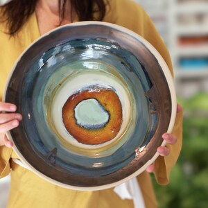 Op de afbeelding: Een handgemaakte keramische kom met een uniek, kleurrijk ontwerp. De kom heeft concentrische cirkels van kleur, waaronder bruin, blauw, groen en oranje, met een lichtgekleurde rand. De kom wordt vastgehouden door een persoon die een geel kledingstuk draagt.