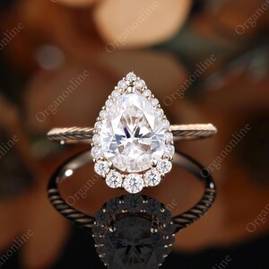 以下が含まれることがあります： ローズゴールドの婚約指輪。 梨型のダイヤモンドが中央にあり、小さな石のハローで囲まれています。 バンドはねじれたロープのデザインです。