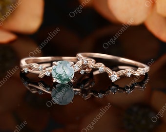 May include: A rose gold ring set featuring a round, green moss agate engagement ring and a matching curved wedding band. Both rings have delicate vine and diamond accents, reflecting light against a dark background.