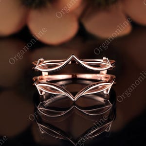 May include: A rose gold ring guard featuring a double-band design with pointed, chevron-like peaks. The metal has a polished finish, and the structure consists of two connected bands that create a symmetrical, open frame for another ring.