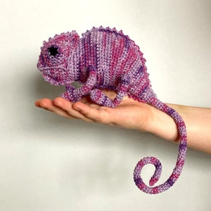 Op de afbeelding: Een gehaakt speelgoedchameleon in roze en paarse tinten. De kameleon heeft een gekrulde staart en wordt in de hand van iemand vastgehouden.