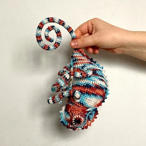 Peut inclure: Un jouet en crochet en forme de caméléon avec une queue enroulée. Le caméléon est fait de fil bleu, blanc et rouge.