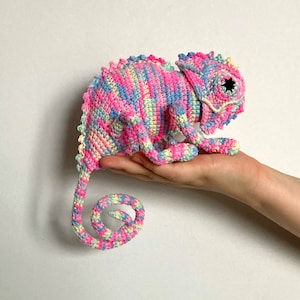 Puede incluir: Un juguete de camaleón de crochet con un patrón de rayas rosa, azul y amarillo. El camaleón tiene una cola rizada y un ojo negro.