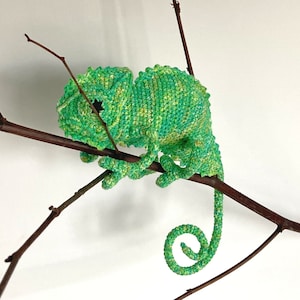 May include: A green crocheted chameleon with a curled tail, perched on a brown branch.