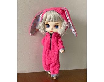 May include: A pink plush bunny onesie with a white hood and a zipper. The onesie has a white and blue patterned trim. The doll has white hair and blue eyes.