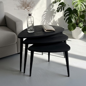 May include: A trio of black nesting tables, each with a triangular top and thin legs. The tables are arranged in a nested formation. A clear glass vase and a book rest on the top table. A grey sofa and a potted plant are in the background.