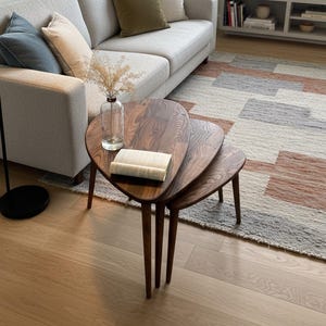 May include: A set of three wooden nesting tables with a dark wood finish. The tables have a unique, rounded triangular shape and are arranged in a tiered configuration. A book rests on the top table. The tables are in a living room setting.