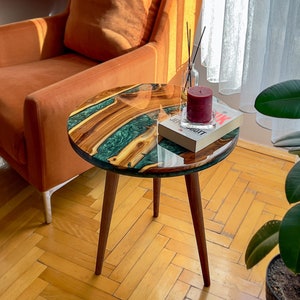 May include: A small, round side table with a wooden top featuring green resin inlay. The table has three angled wooden legs. A red candle and a book rest on the table. The table's diameter is approximately 40 cm.