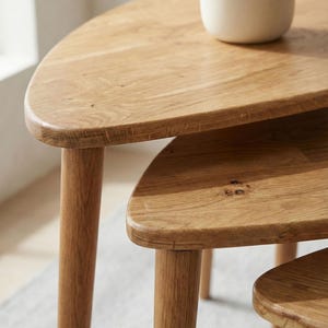 Close-up detail of a handcrafted solid wood nesting table set highlighting smooth rounded edge profiles and refined joinery. Natural oak grain patterns with subtle knots and tonal variations are enhanced by soft daylight. The layered design showcases the stepped arrangement of three tables with tapered cylindrical legs, emphasizing Scandinavian minimalist style and space-saving functionality within a calm neutral interior setting.