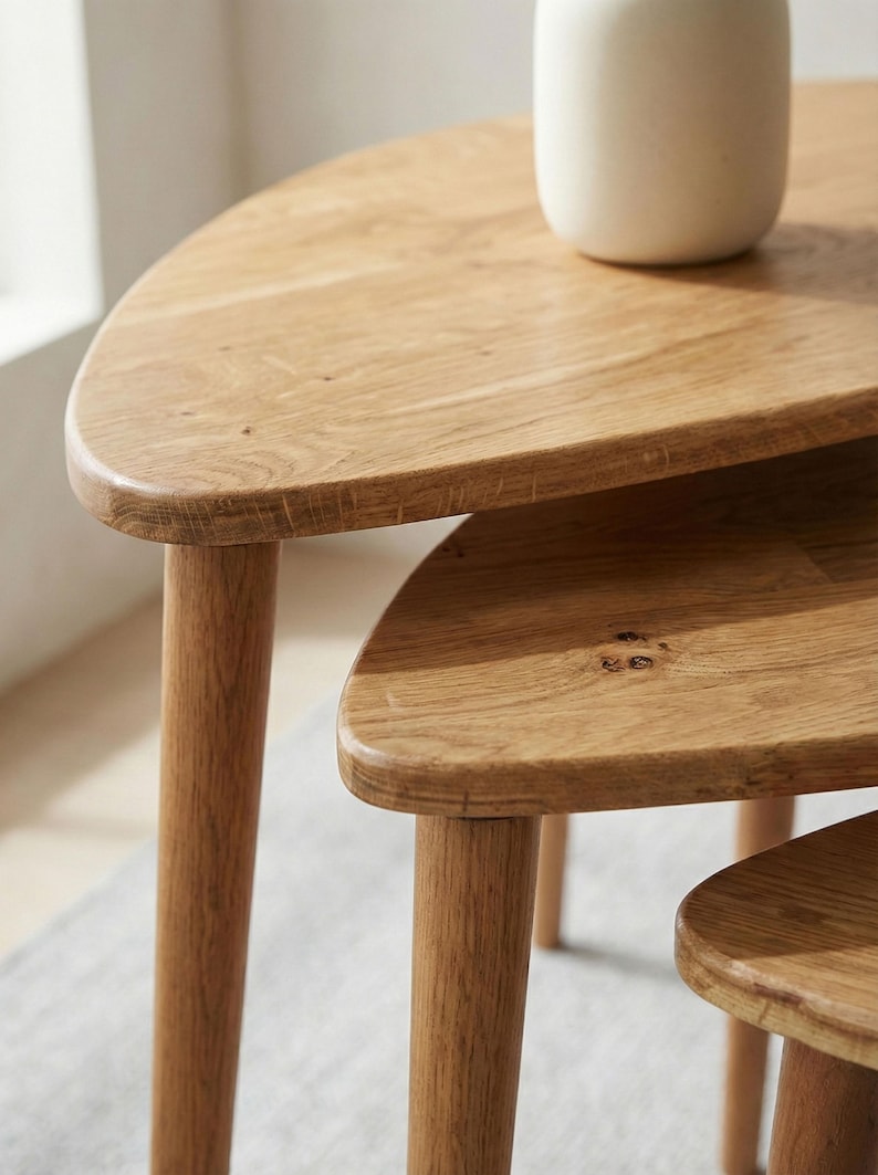 Close-up detail of a handcrafted solid wood nesting table set highlighting smooth rounded edge profiles and refined joinery. Natural oak grain patterns with subtle knots and tonal variations are enhanced by soft daylight. The layered design showcases the stepped arrangement of three tables with tapered cylindrical legs, emphasizing Scandinavian minimalist style and space-saving functionality within a calm neutral interior setting.