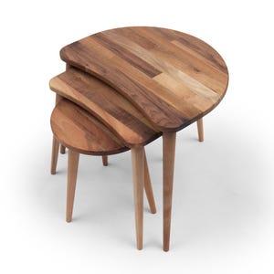 May include: A set of three nesting tables with a natural wood finish. The tables have a unique, curved design and are supported by tapered legs. The largest table is approximately 24 inches in diameter.