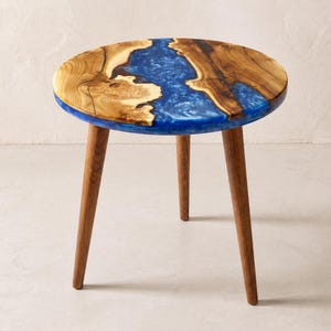 May include: A round side table featuring a natural wood top with a central river-like inlay of translucent blue resin. The table is supported by three tapered wooden legs that extend outward from the center, providing a stable base for the piece.