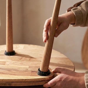 Close-up of assembly process showing a handcrafted solid wood nesting table with a tapered leg being securely fitted into the tabletop. The smooth oak surface reveals natural grain patterns and warm tones, while the leg connection detail highlights sturdy joinery and precision craftsmanship. Hands-on interaction emphasizes easy setup, durable construction, and the quality of real wood materials used in the design.