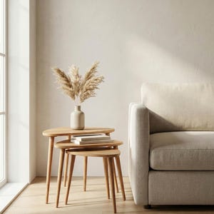Lifestyle scene of a handcrafted solid wood nesting table set placed beside a neutral fabric sofa in a bright minimalist interior. Three organically shaped tables with smooth rounded edges and tapered legs are stacked together, showcasing space-saving functionality. Natural oak grain and warm tones are enhanced by soft daylight from the window, while a ceramic vase with dried pampas grass and stacked books add a calm, modern styling touch.
