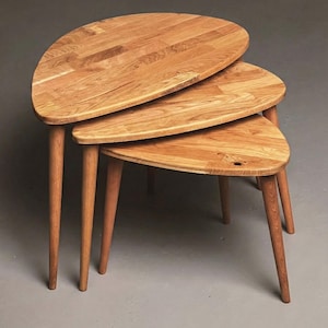 Angled studio view of a handcrafted solid oak nesting table set featuring three organically shaped tabletops with smooth rounded edges. The layered arrangement highlights the space-saving nesting function, while slender tapered legs add a refined mid-century modern character. Natural oak grain, knots, and warm tonal variations are clearly visible, set against a clean neutral backdrop to emphasize material quality and craftsmanship.