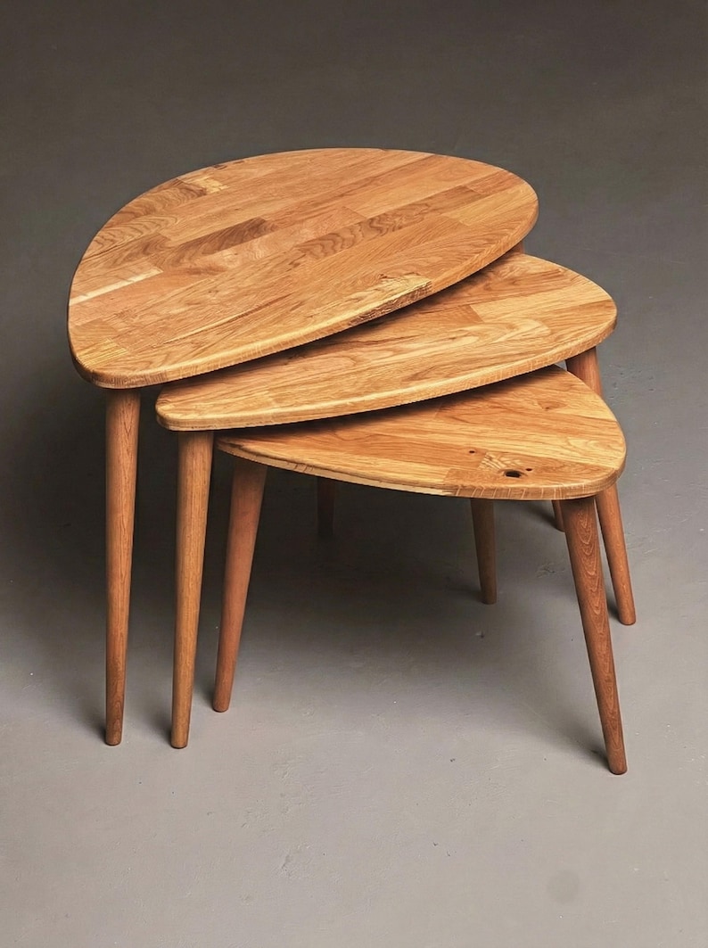 Angled studio view of a handcrafted solid oak nesting table set featuring three organically shaped tabletops with smooth rounded edges. The layered arrangement highlights the space-saving nesting function, while slender tapered legs add a refined mid-century modern character. Natural oak grain, knots, and warm tonal variations are clearly visible, set against a clean neutral backdrop to emphasize material quality and craftsmanship.