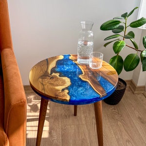 May include: A round side table with a wooden top and blue resin river design. The table has three wooden legs and is topped with a clear glass carafe and a drinking glass. The table is a unique piece of furniture.