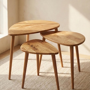 Angled perspective of a handcrafted solid wood nesting table set with three organically shaped tabletops in warm oak tones. The staggered heights and overlapping design highlight practical space-saving use. Smooth rounded edges and tapered legs emphasize Scandinavian minimalist style. Natural wood grain, knots, and tonal variations are clearly visible, enhanced by soft sunlight in a neutral interior, creating a clean and modern aesthetic.