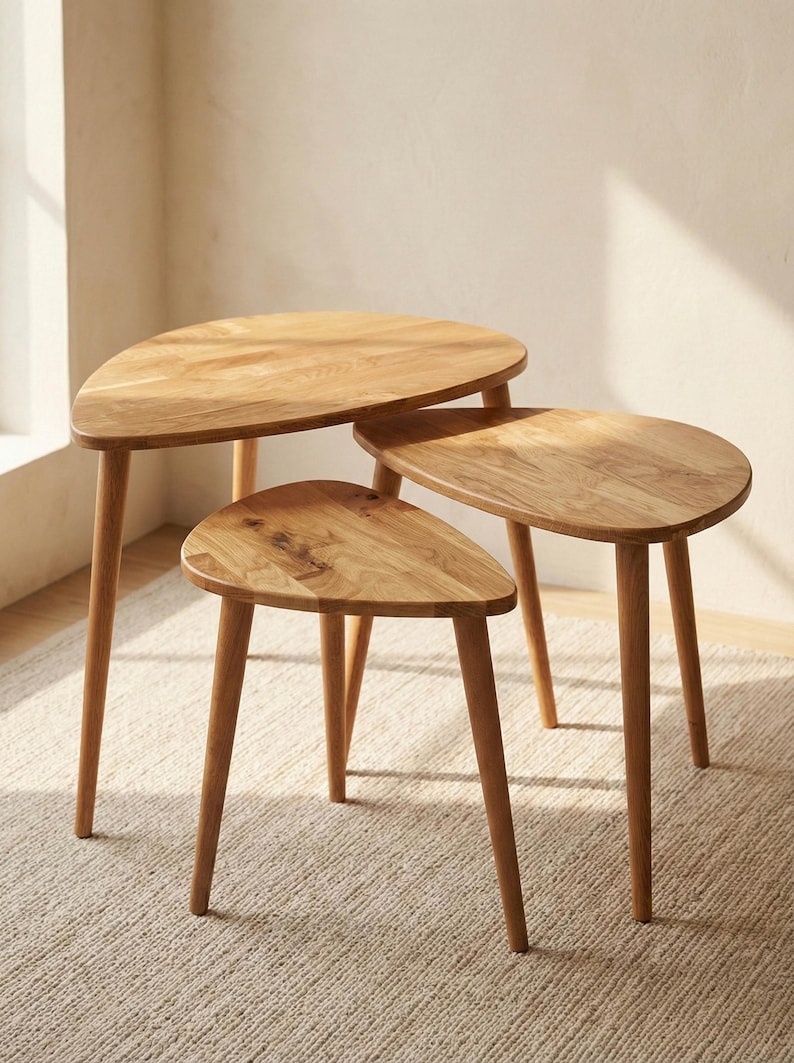 Angled perspective of a handcrafted solid wood nesting table set with three organically shaped tabletops in warm oak tones. The staggered heights and overlapping design highlight practical space-saving use. Smooth rounded edges and tapered legs emphasize Scandinavian minimalist style. Natural wood grain, knots, and tonal variations are clearly visible, enhanced by soft sunlight in a neutral interior, creating a clean and modern aesthetic.