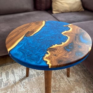May include: Round wooden coffee table with a blue resin river design. The table features a walnut wood grain and three wooden legs. The blue resin creates a flowing river effect across the tabletop, adding a unique aesthetic to the furniture.