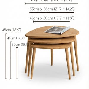 Product dimension display of a handcrafted solid wood nesting table set featuring three organically shaped tables with smooth rounded edges and tapered legs. The image shows size variations with labeled measurements in both centimeters and inches, highlighting width and height differences for each piece. Natural oak grain and warm tones are visible, while the stacked arrangement emphasizes practical nesting functionality and space-saving design.