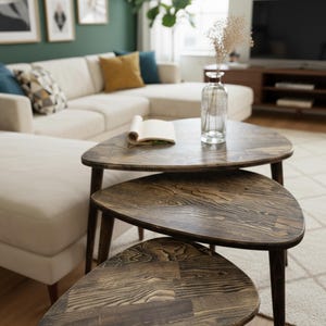 May include: Set of three nesting tables with a dark wood grain finish. The tables have a triangular shape with rounded edges and dark brown legs. A clear glass vase with dried flowers sits on the top table. The tables are in a living room setting.
