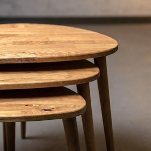 Close-up side view of a handcrafted solid oak nesting table set showing three layered tabletops with smooth rounded edges. The staggered heights highlight the functional nesting design, while slim tapered legs create a clean, mid-century inspired look. Rich oak grain, natural knots, and warm brown tones are emphasized under soft lighting, showcasing authentic wood texture and refined craftsmanship.