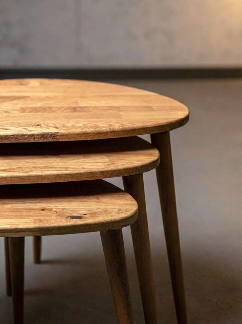Close-up side view of a handcrafted solid oak nesting table set showing three layered tabletops with smooth rounded edges. The staggered heights highlight the functional nesting design, while slim tapered legs create a clean, mid-century inspired look. Rich oak grain, natural knots, and warm brown tones are emphasized under soft lighting, showcasing authentic wood texture and refined craftsmanship.
