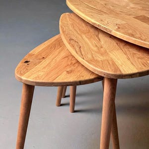 Close-up angled view of a handcrafted solid oak nesting table set featuring three organically shaped tabletops with smooth rounded edges. The layered arrangement highlights the space-saving nesting design, while tapered wooden legs add a refined mid-century touch. Natural oak grain, knots, and tonal variations are clearly visible, emphasizing authentic wood character and durable craftsmanship against a clean, neutral background.