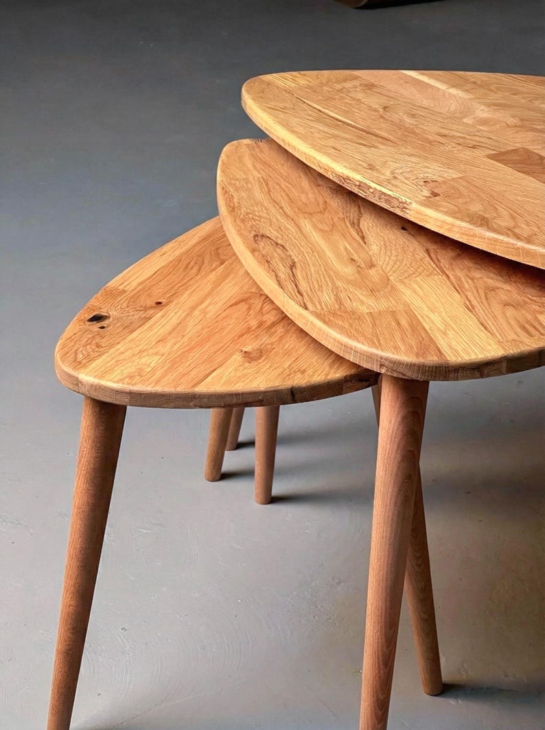 Close-up angled view of a handcrafted solid oak nesting table set featuring three organically shaped tabletops with smooth rounded edges. The layered arrangement highlights the space-saving nesting design, while tapered wooden legs add a refined mid-century touch. Natural oak grain, knots, and tonal variations are clearly visible, emphasizing authentic wood character and durable craftsmanship against a clean, neutral background.