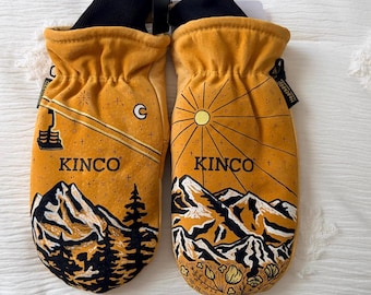 May include: A pair of mustard yellow mittens with black cuffs. Each mitten features a mountain landscape design with the word "KINCO" printed in black. One mitten has a sunburst design, the other has a moon and stars.